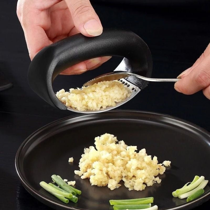 Hand using a garlic press to crush garlic onto a black plate with green onions.